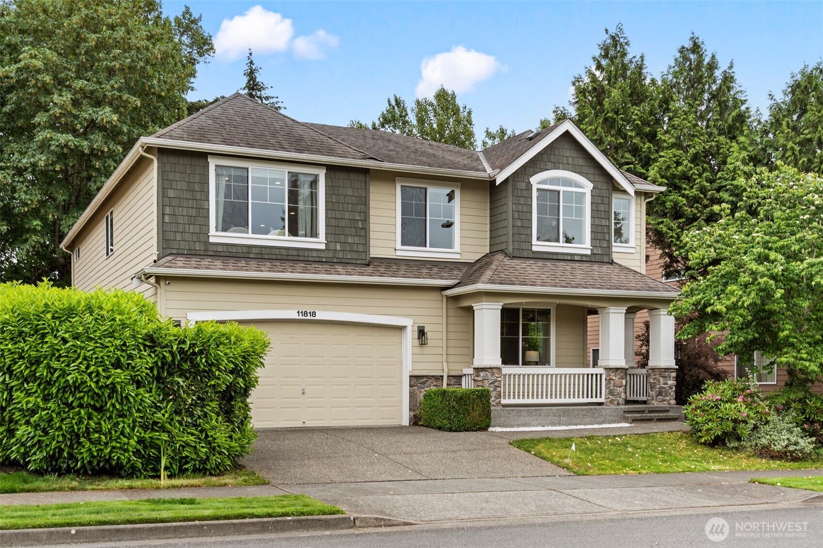 Two-story beige house with gray shingle accents, a covered porch, and mature landscaping.