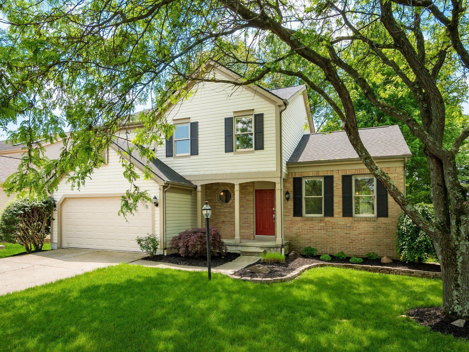 Two-story house with cream siding, brick accents, red front door, and attached garage beneath mature trees.