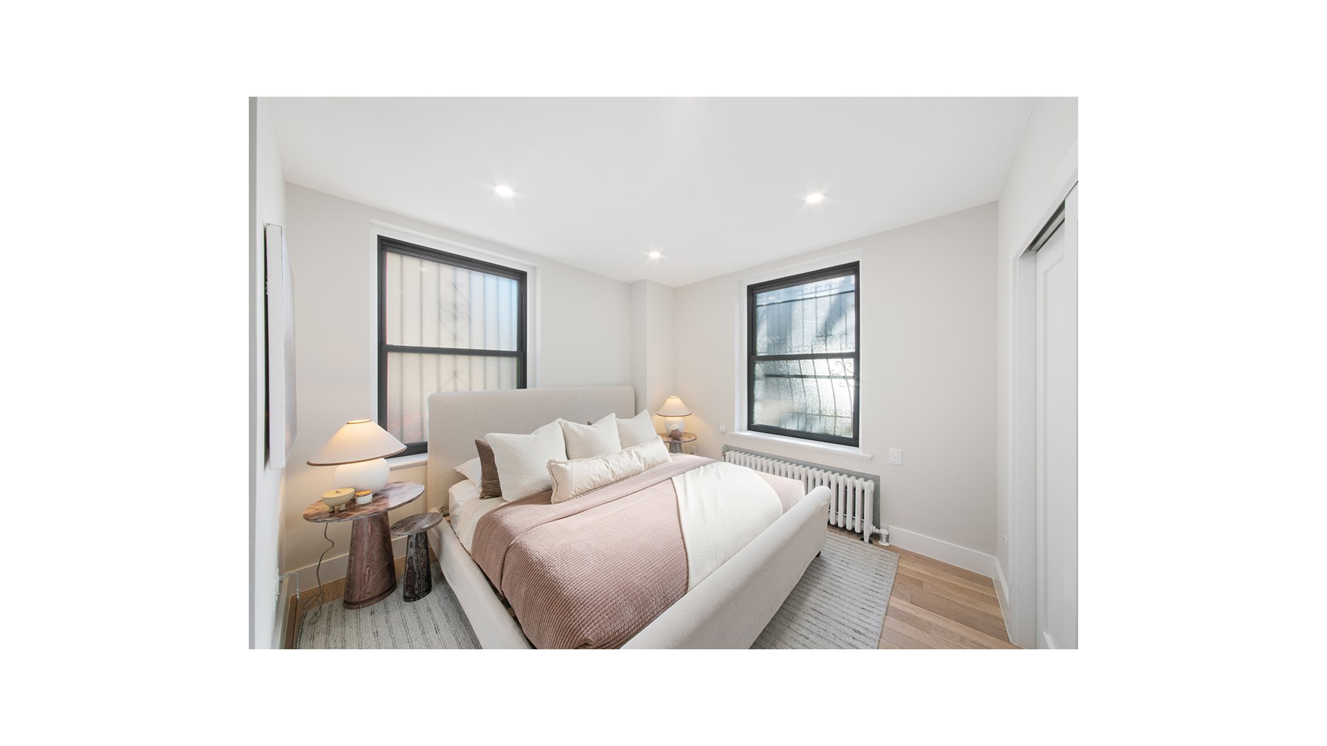 Staged bedroom with a white bed, two large windows, and a radiator.