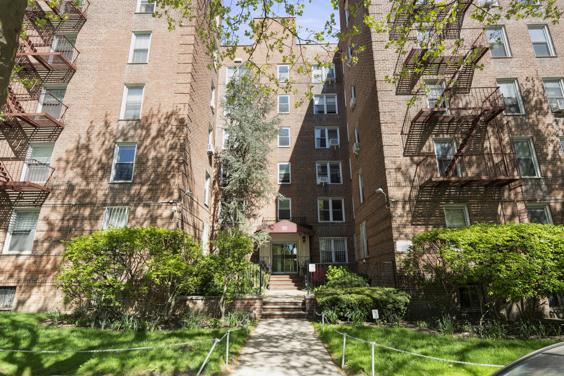 Brick apartment building entrance with fire escapes and landscaped front garden.