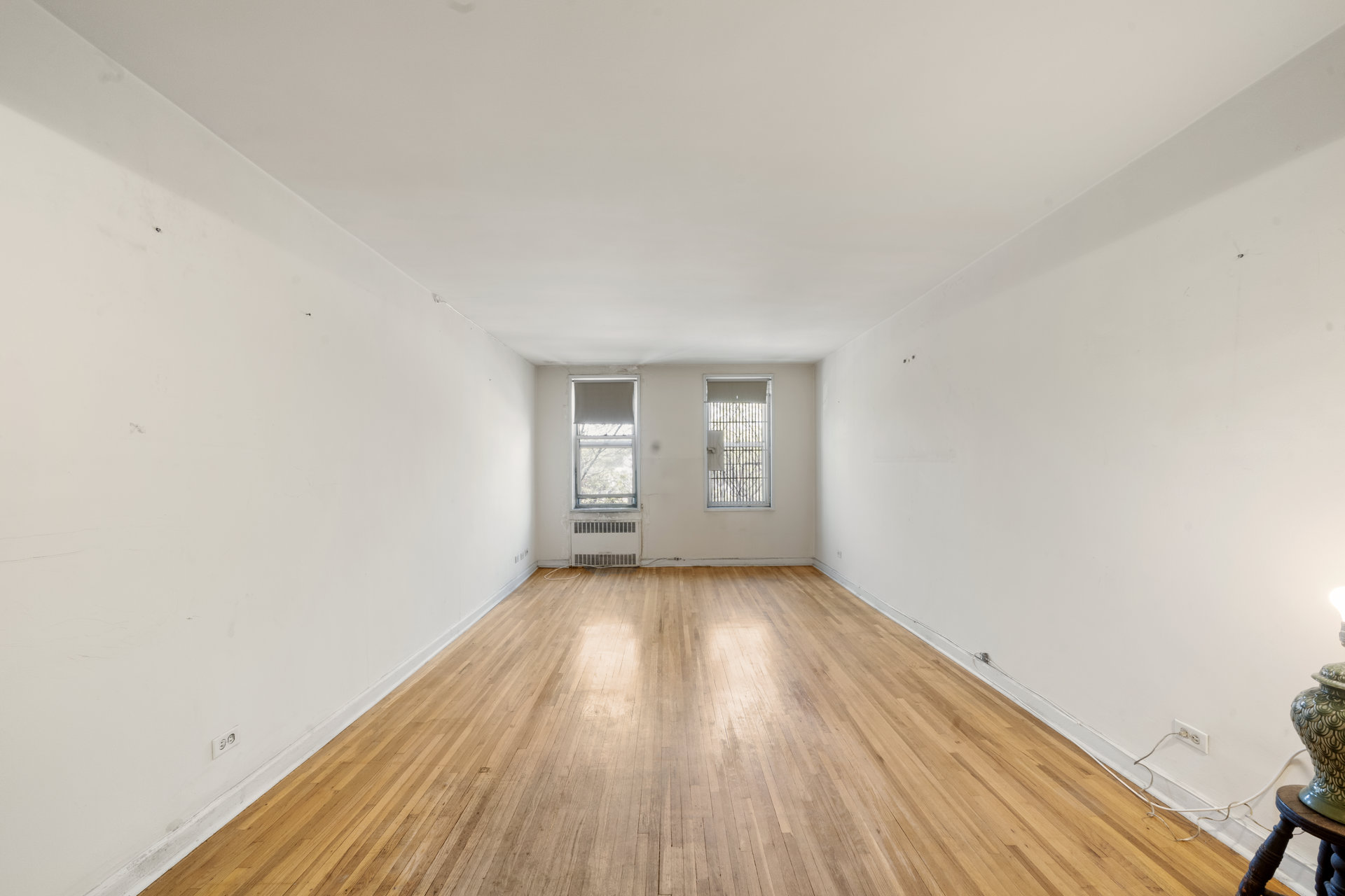 Long, empty room with hardwood floors and two windows at the far end.
