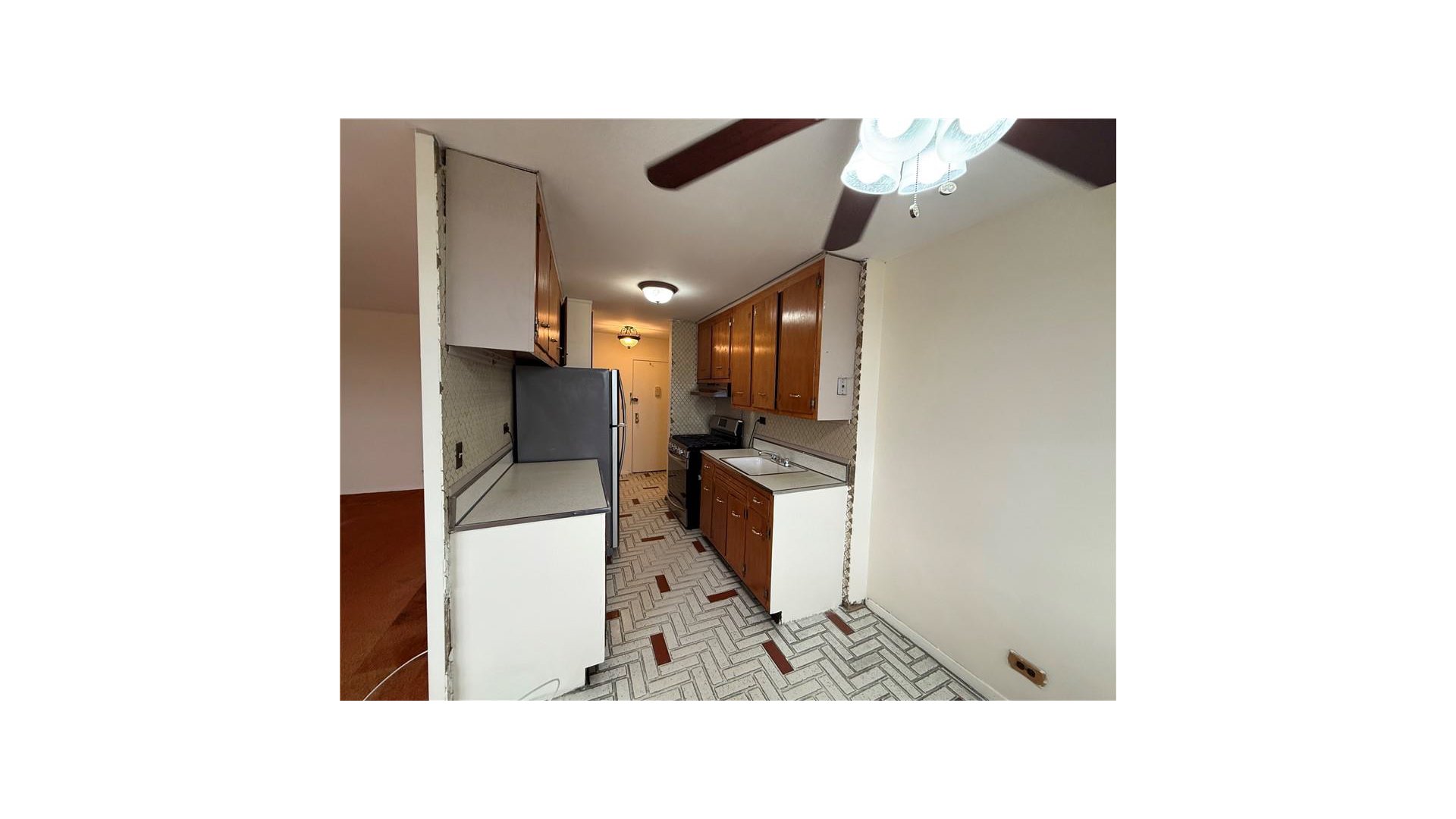 Narrow kitchen with wood cabinets, patterned tile floor, refrigerator, and a ceiling fan.