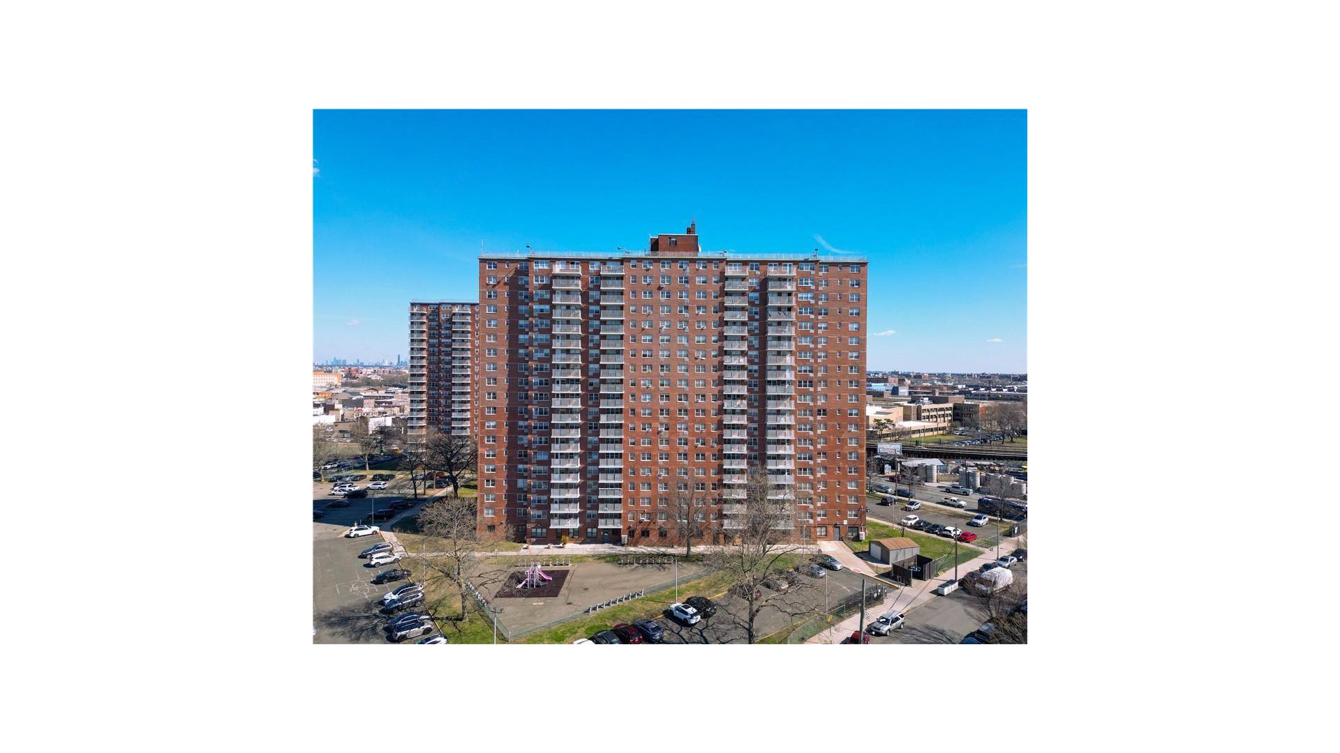 Red-brick apartment tower with rows of balconies overlooking a small playground and parking areas.