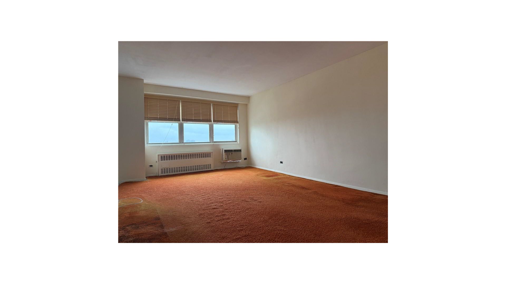 Vacant living room with wall-to-wall rust-colored carpet and wide windows with blinds.