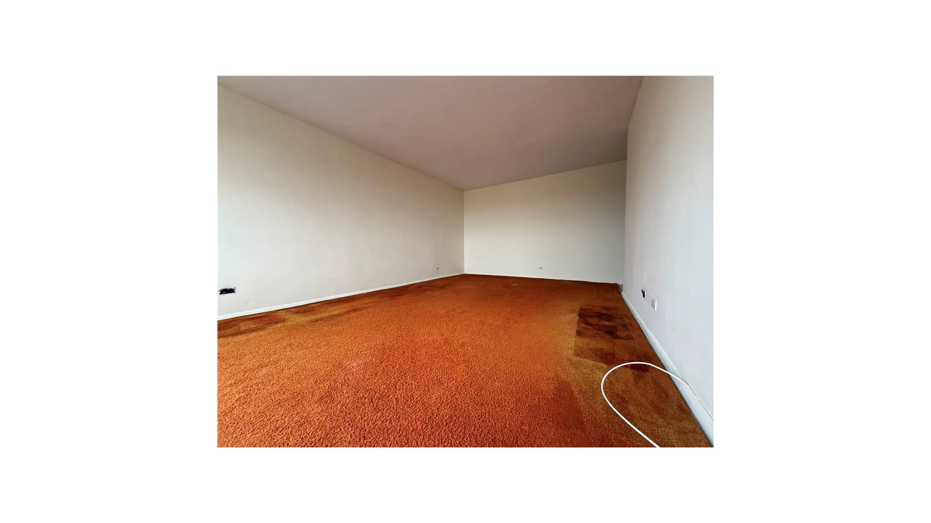 Empty room with orange-brown carpet, white walls, and an outlined area on the floor.