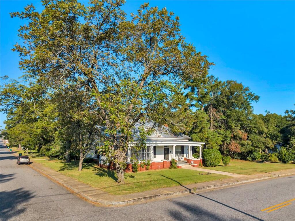 White single-story house with a red brick foundation and covered porch beneath a large tree.