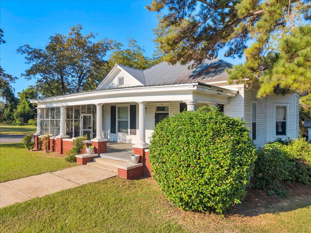 White-sided house with a broad columned porch, red brick supports, and mature landscaping.