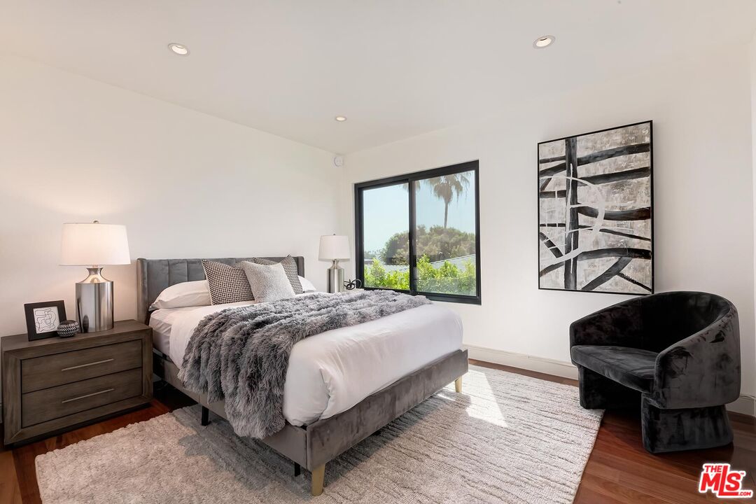 Staged bedroom with a gray upholstered bed, black accent chair, and large window overlooking greenery.