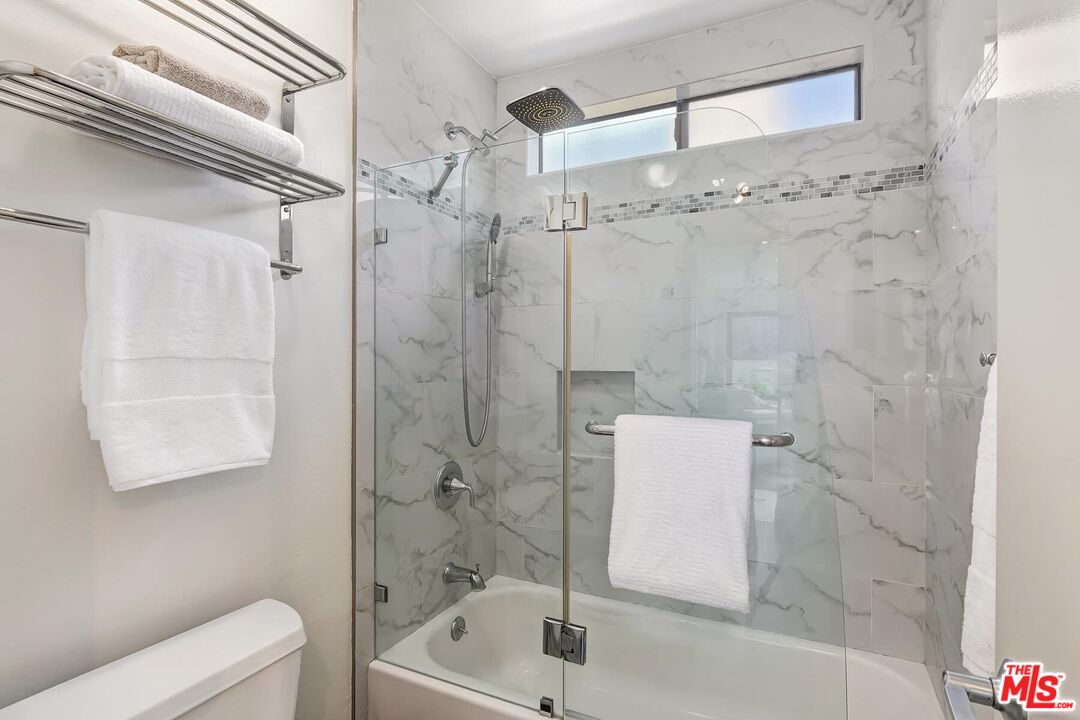 Bathroom with a glass-enclosed tub, marble-look tile, rainfall showerhead, and high window.