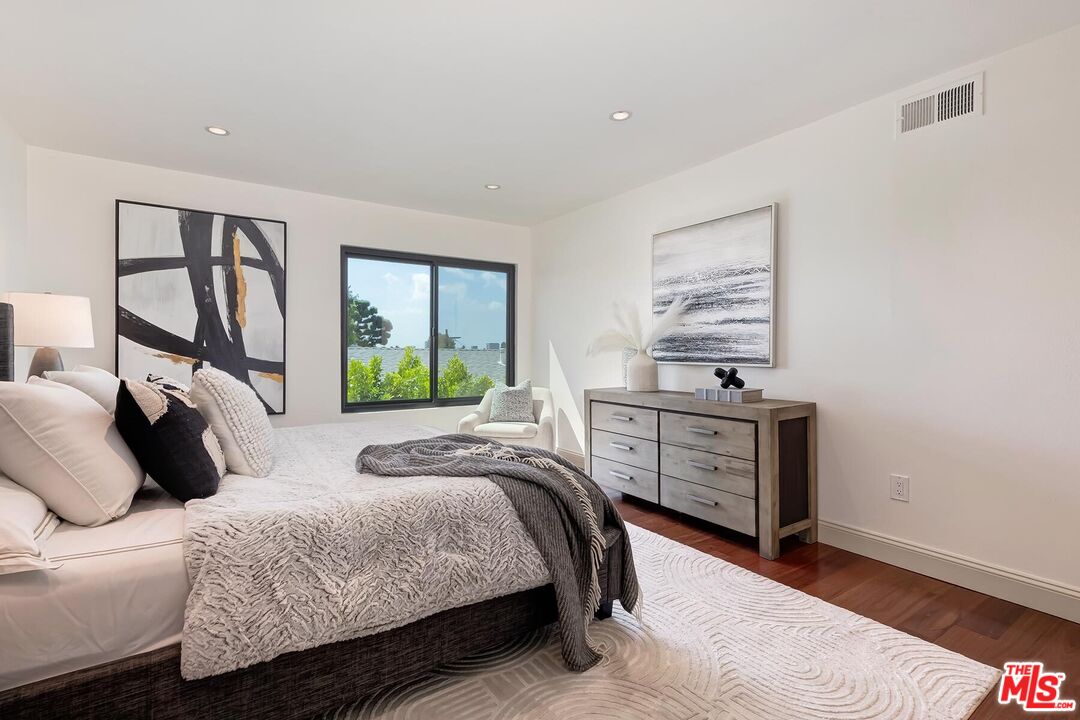 Furnished bedroom with a large bed, abstract art, wood dresser, and window overlooking greenery.