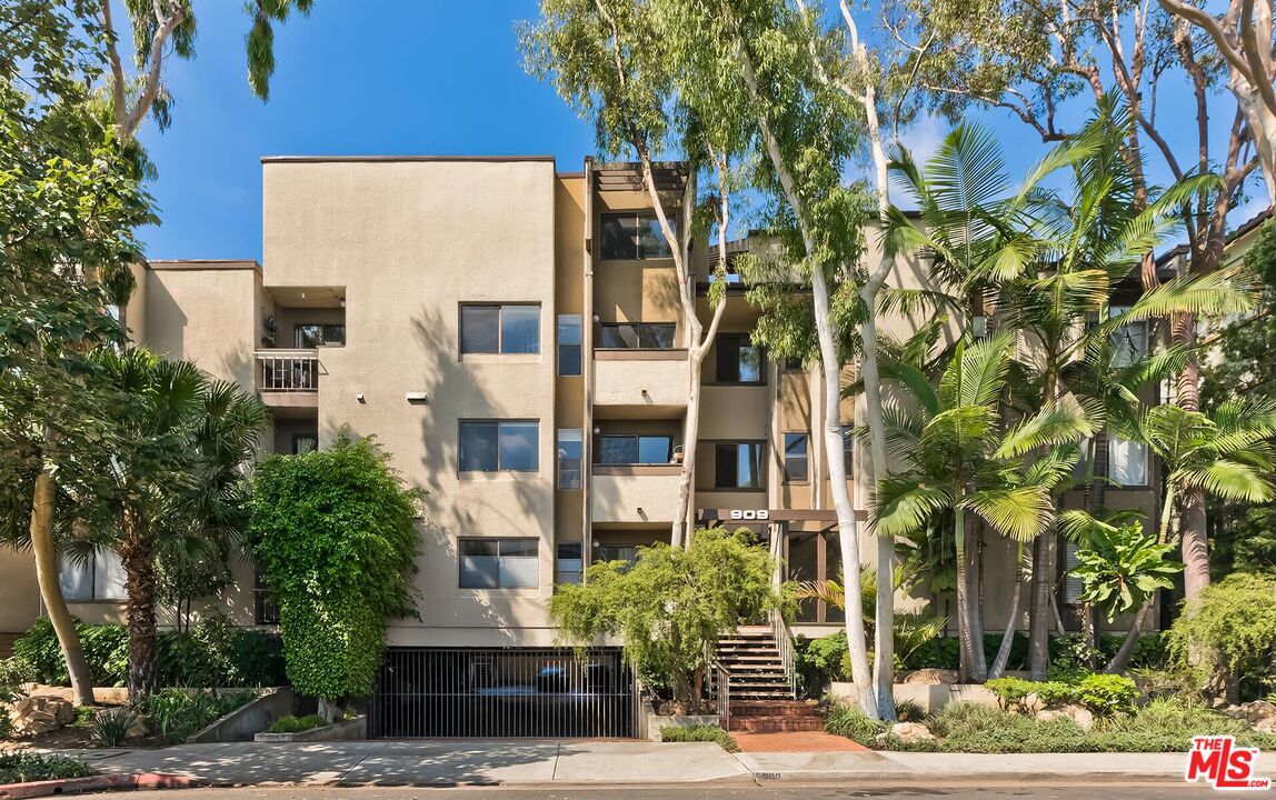 Beige apartment building with balconies, palms, and a gated garage entrance.
