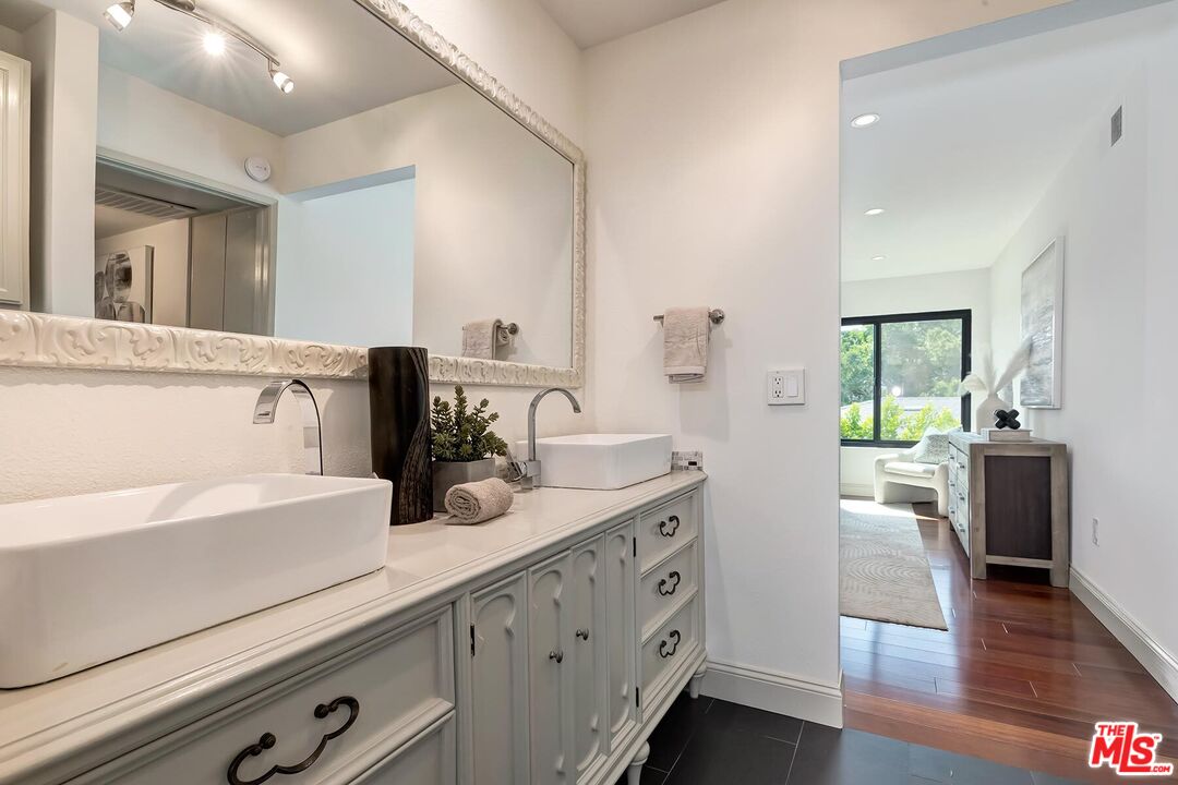 Bathroom with a white double vanity, vessel sinks, ornate mirror, and doorway to a bright bedroom.