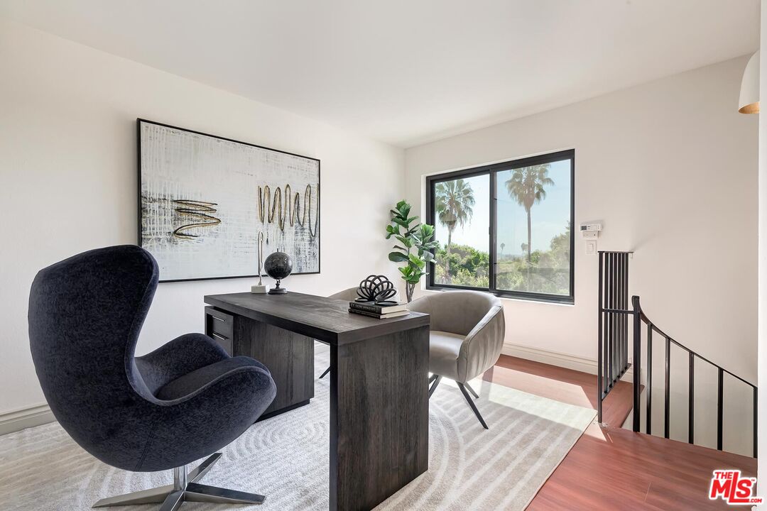 Staged home office with a dark desk, sculptural chairs, and a window overlooking palm trees.