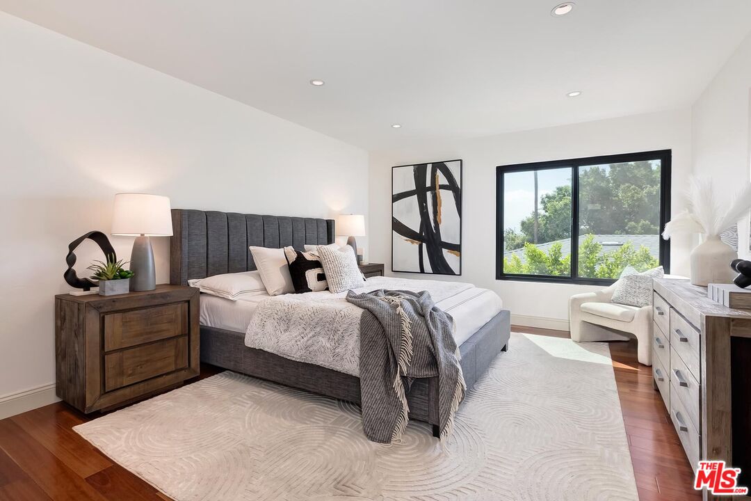 Staged bedroom with a gray upholstered bed, pale patterned rug, and large window overlooking trees.