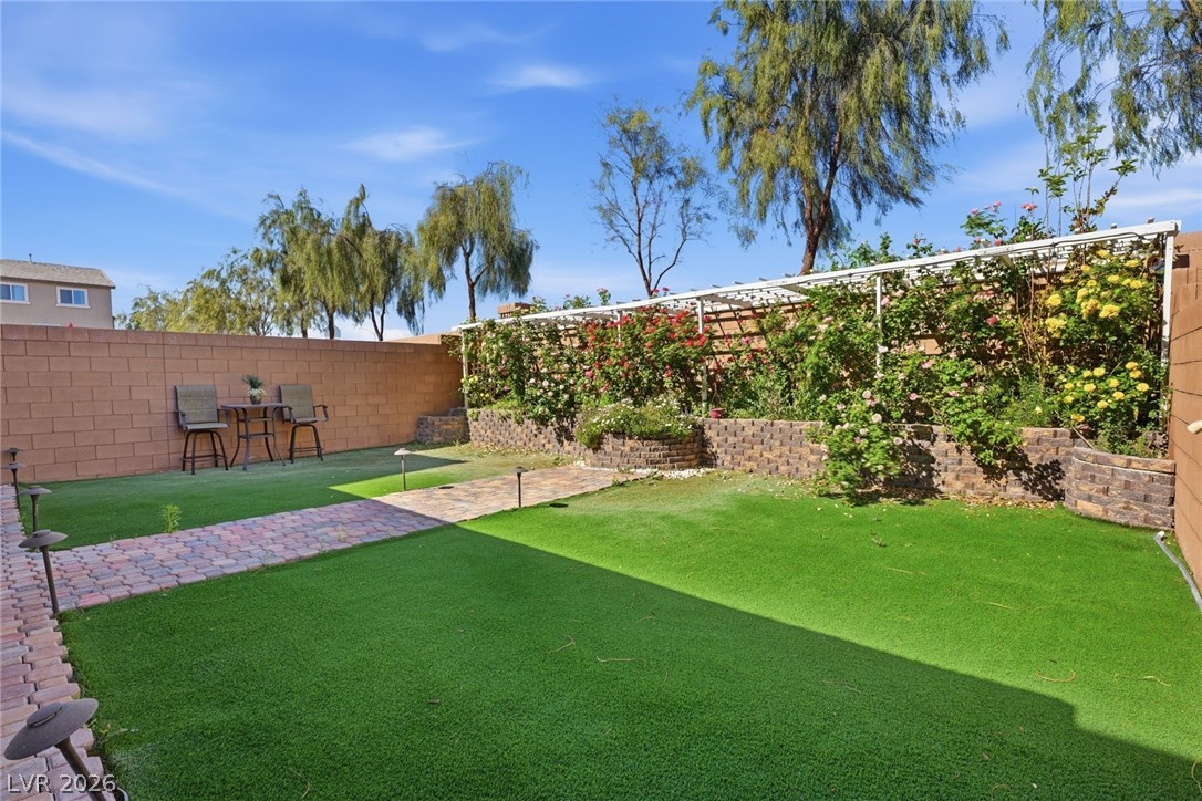 Backyard with artificial turf, a paver path, and flowering vines beneath a pergola.