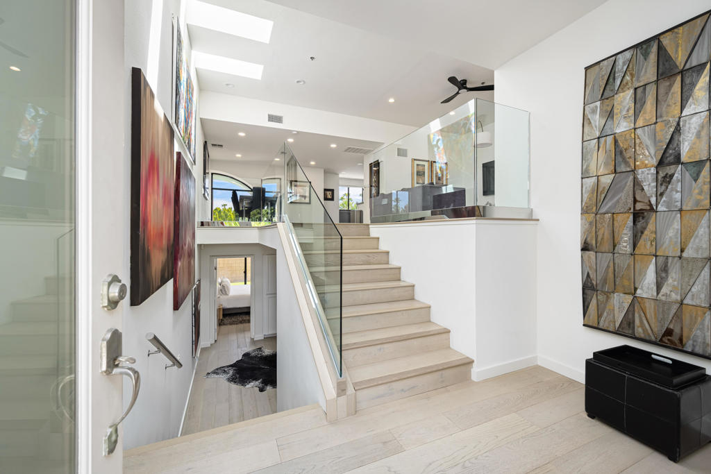 Modern foyer with a pale wood staircase, glass railing, and geometric wall art.