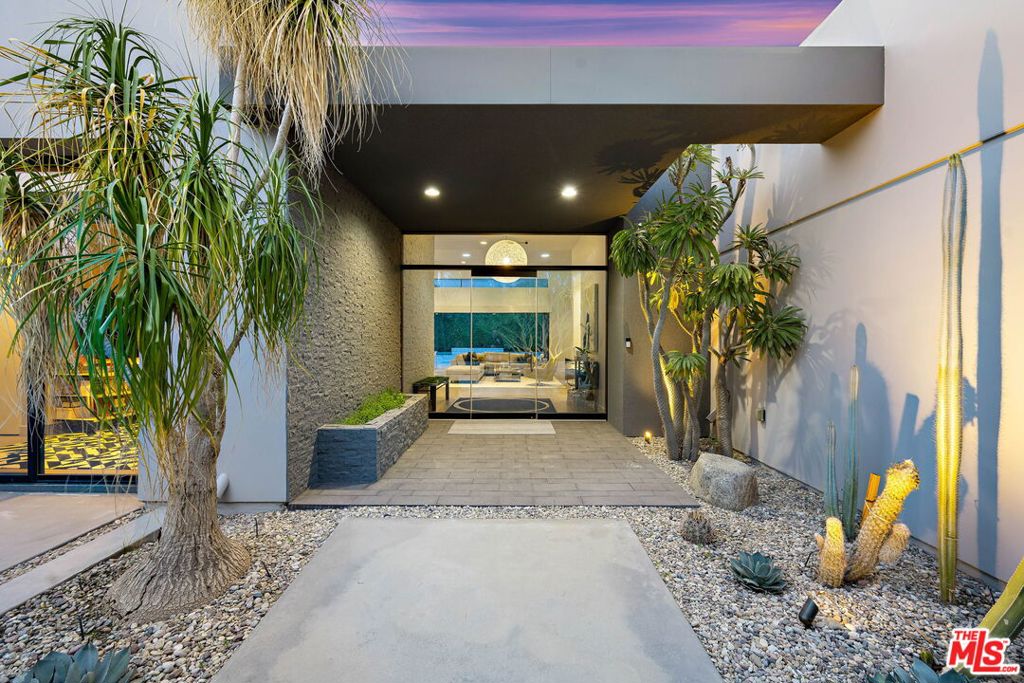 Modern home entrance with glass doors, a covered walkway, and desert landscaping at dusk.