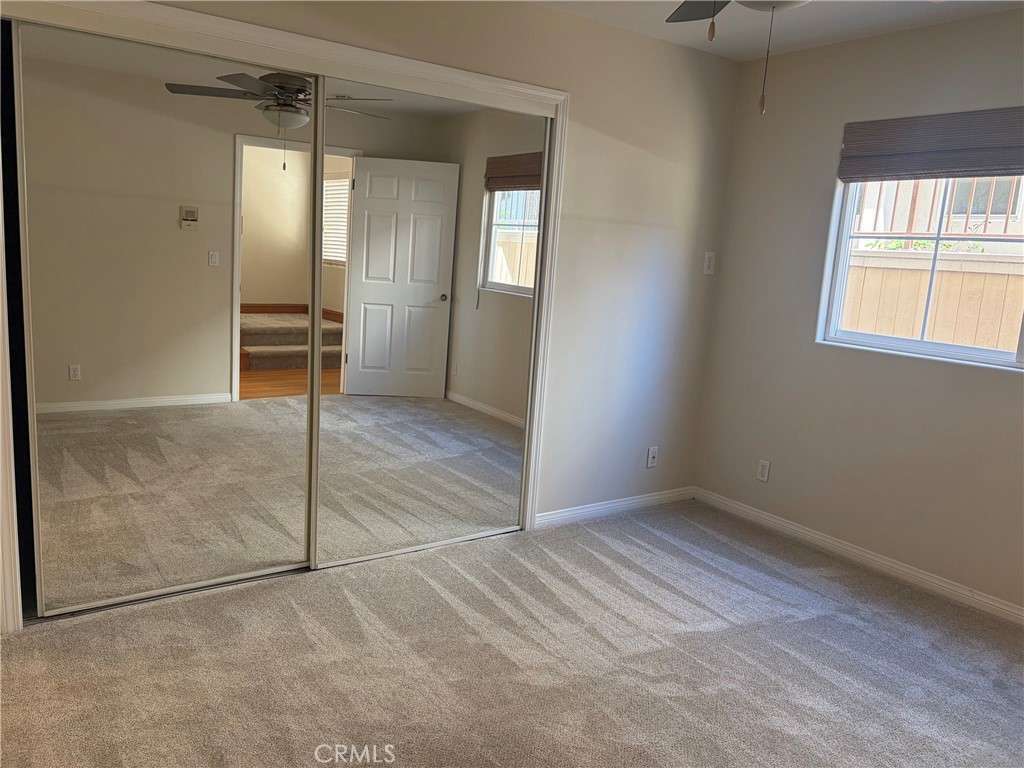Empty carpeted bedroom with mirrored closet doors, a window, and a ceiling fan.