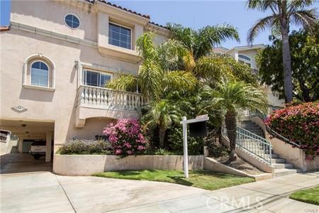 Two-story stucco home with a balcony, garage, exterior stairs, palm trees, and flowering shrubs.