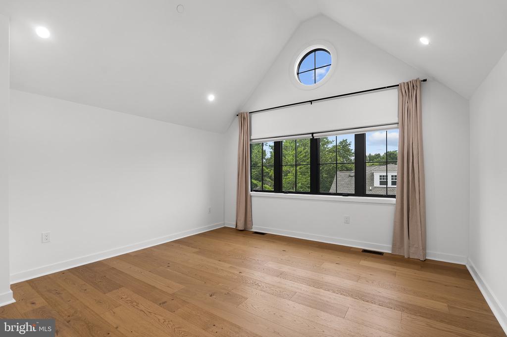 Vacant room with hardwood floors, vaulted ceiling, round window, and wide curtained window.