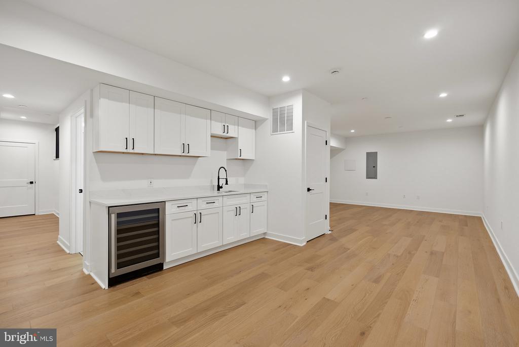 White kitchenette with beverage refrigerator and light wood floors opening into an empty room.