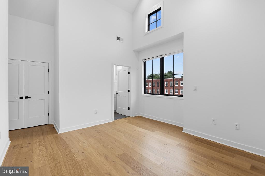 Vacant bedroom with tall ceilings, hardwood floors, and large windows overlooking a brick building.