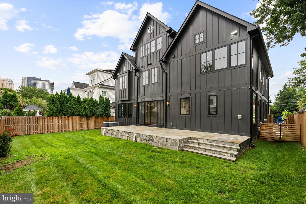 Dark gray multi-story home with large windows, raised stone patio, and fenced backyard lawn.
