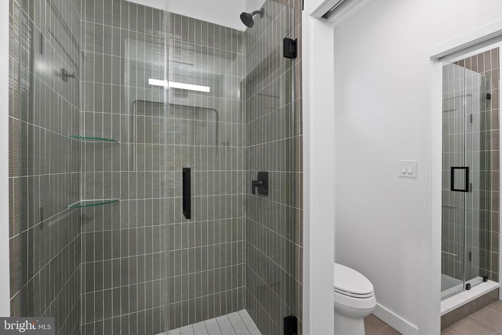 Modern tiled walk-in shower with black fixtures and glass shelves beside a toilet.