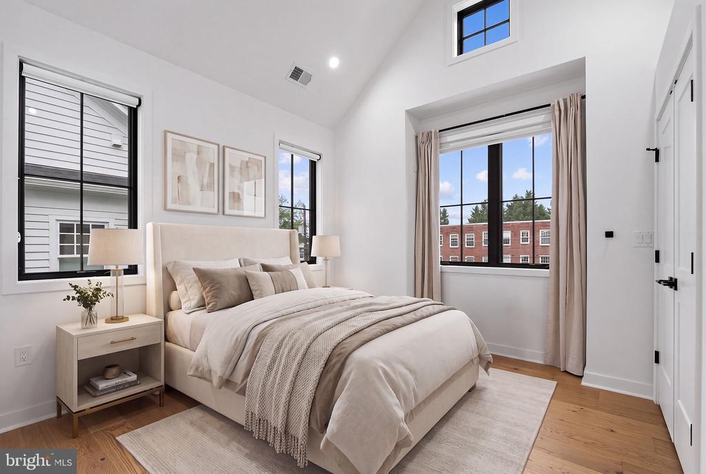 Staged bedroom with a neutral bed, tall black-framed windows, and warm wood floors.