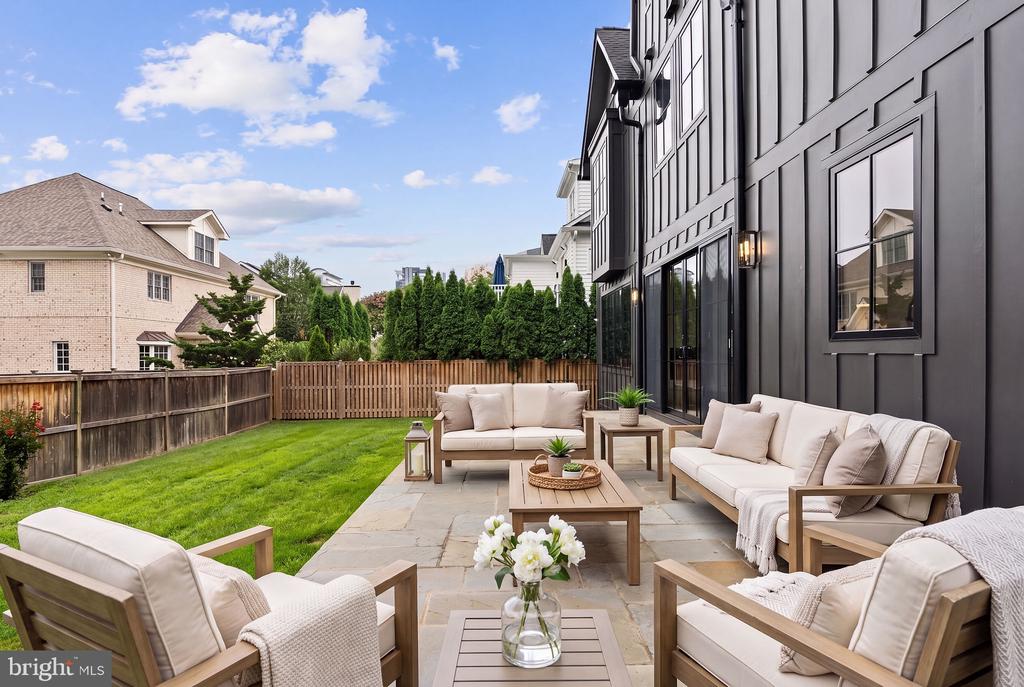 Furnished backyard patio with light-colored seating beside a modern dark-sided house and fenced lawn.