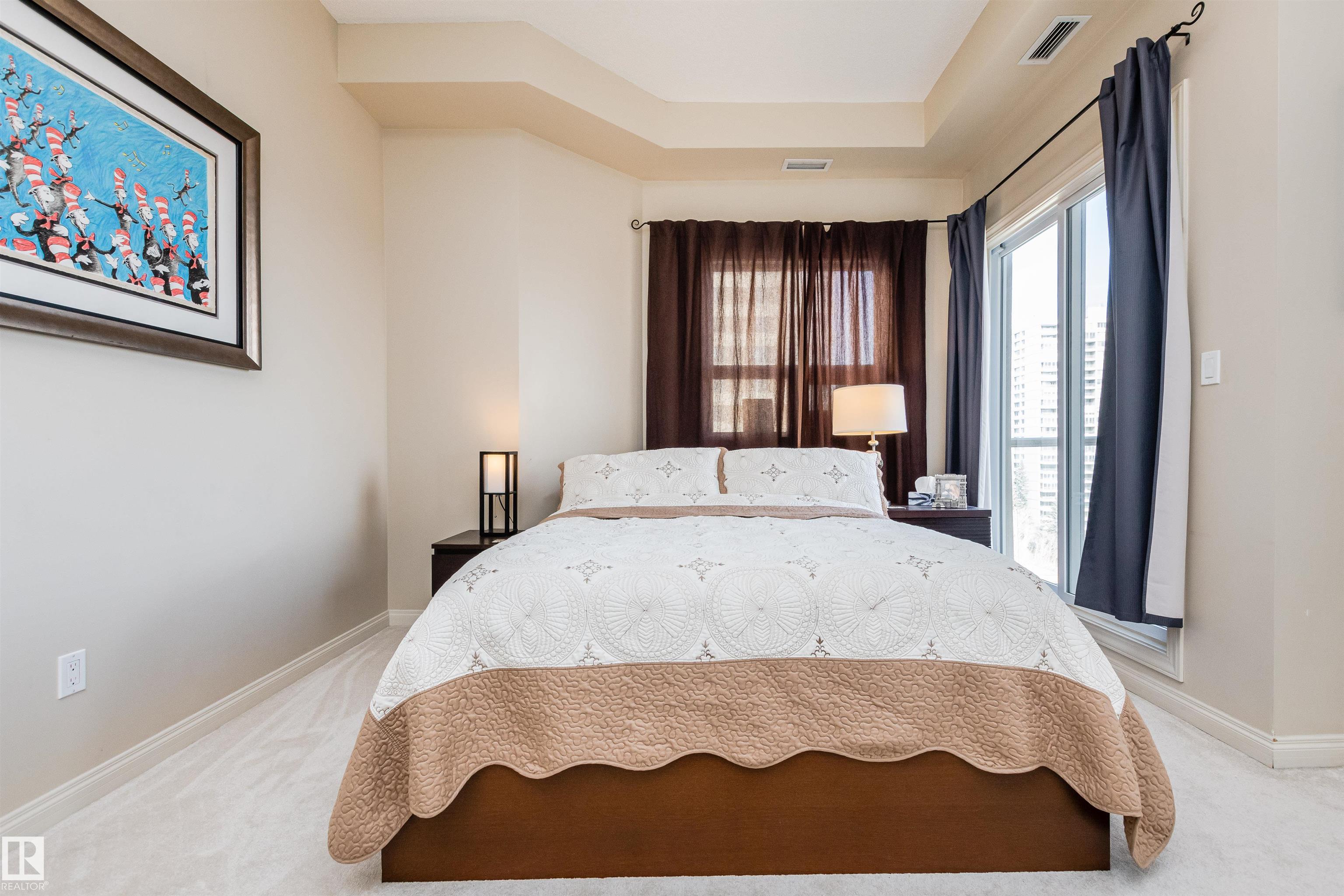 Furnished bedroom with a large patterned bed, dark curtains, and a tall window overlooking nearby buildings.