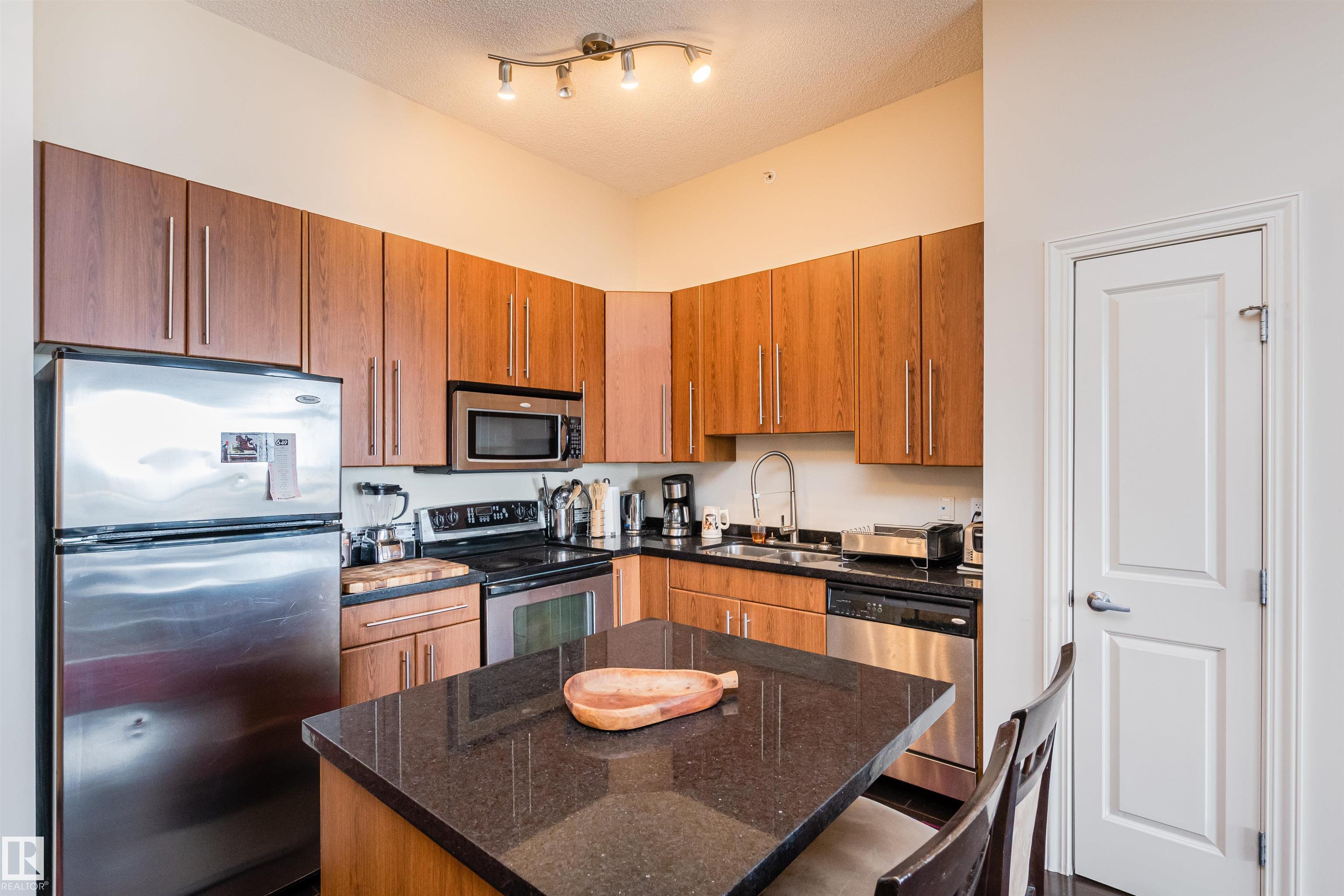 Condo kitchen with warm wood cabinetry, stainless steel appliances, and a dark stone island.