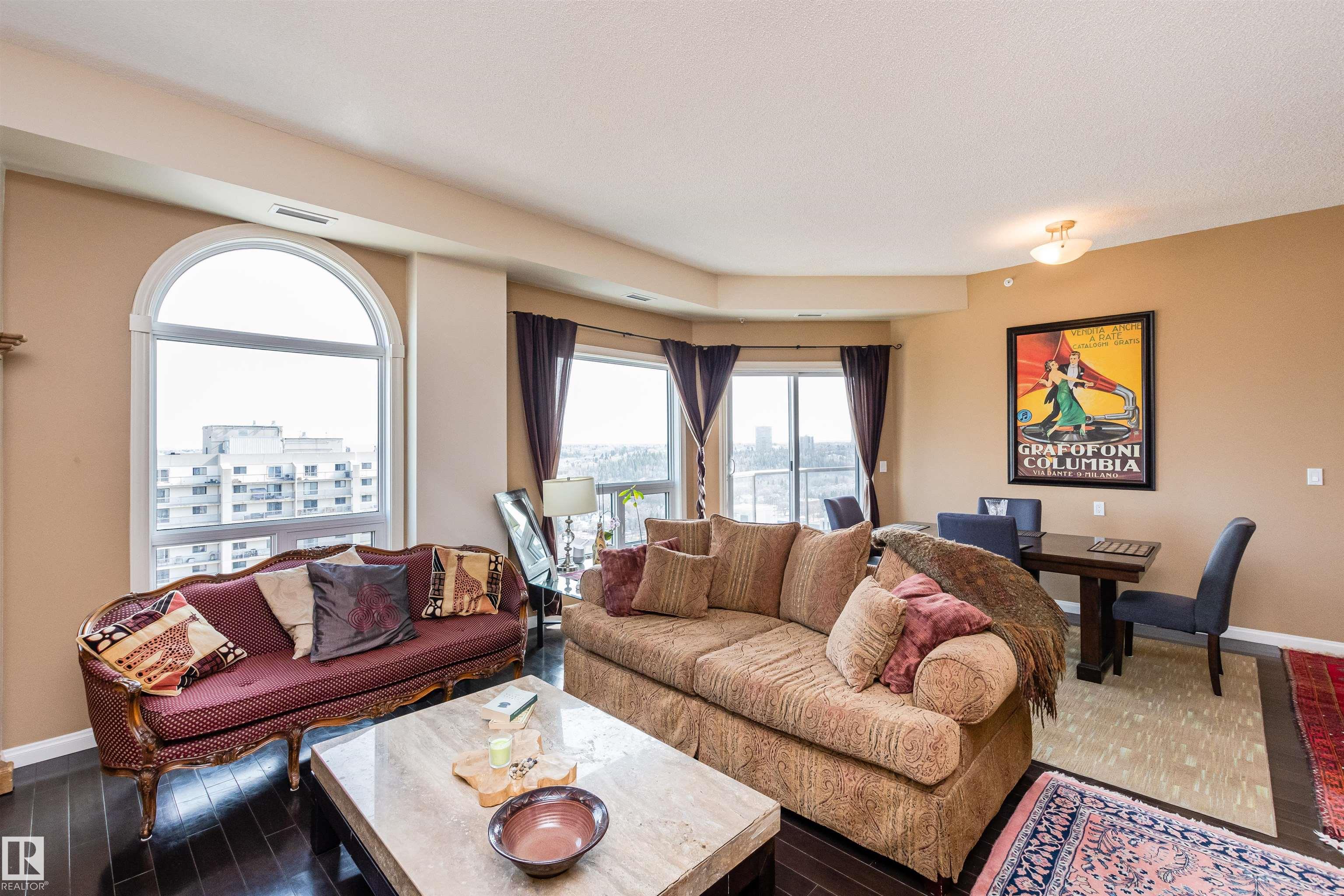 Furnished condo living room with patterned sofas, a marble coffee table, and large windows overlooking nearby buildings.
