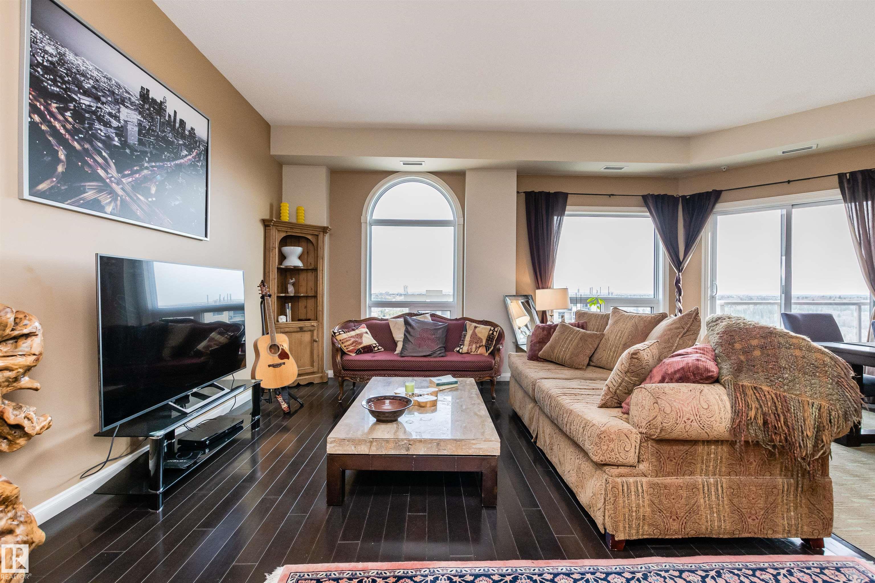 Furnished living room with a beige sectional, dark hardwood floors, and large windows overlooking the city.