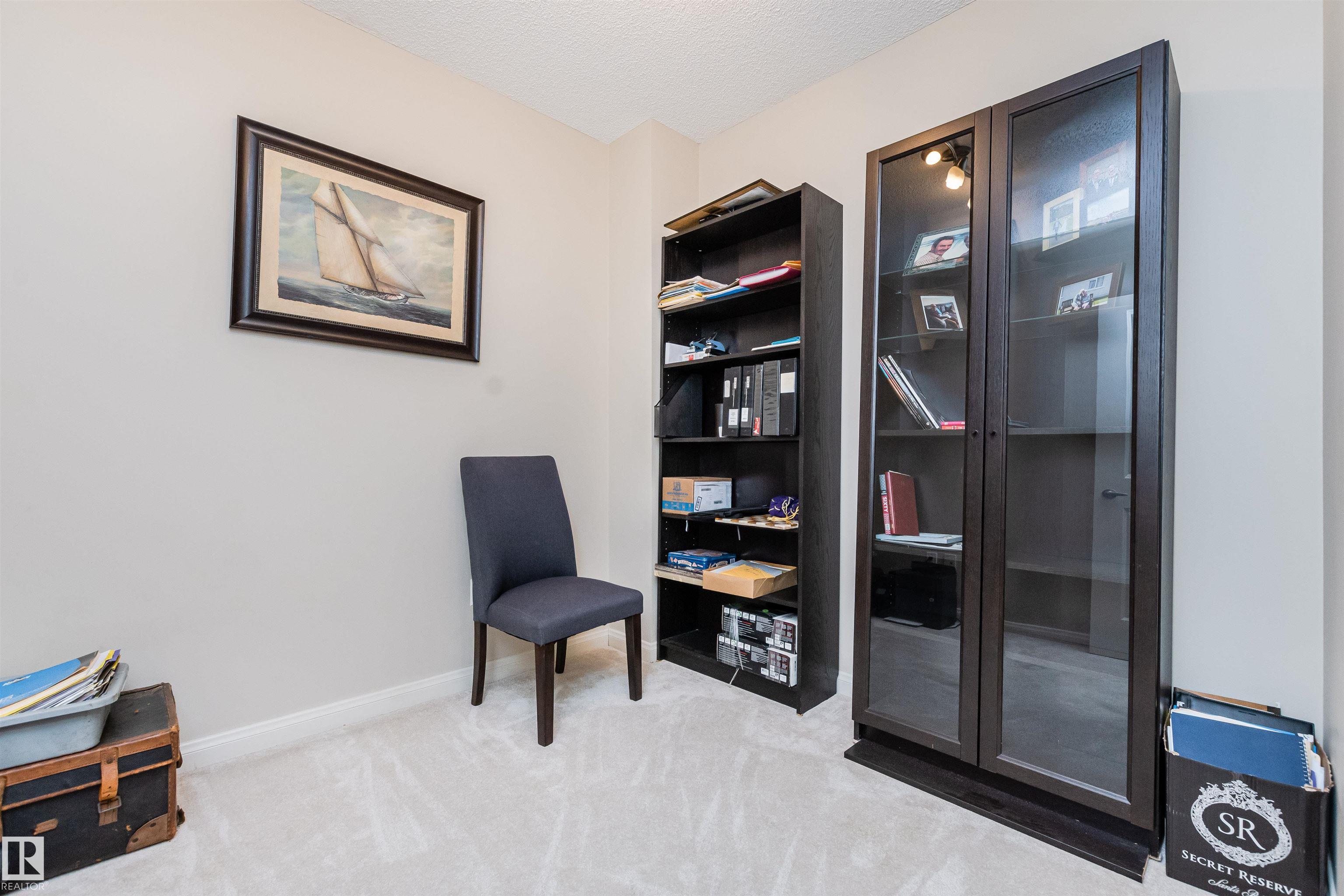Home office with a gray chair, dark shelving and glass-front cabinet, and framed sailboat painting.
