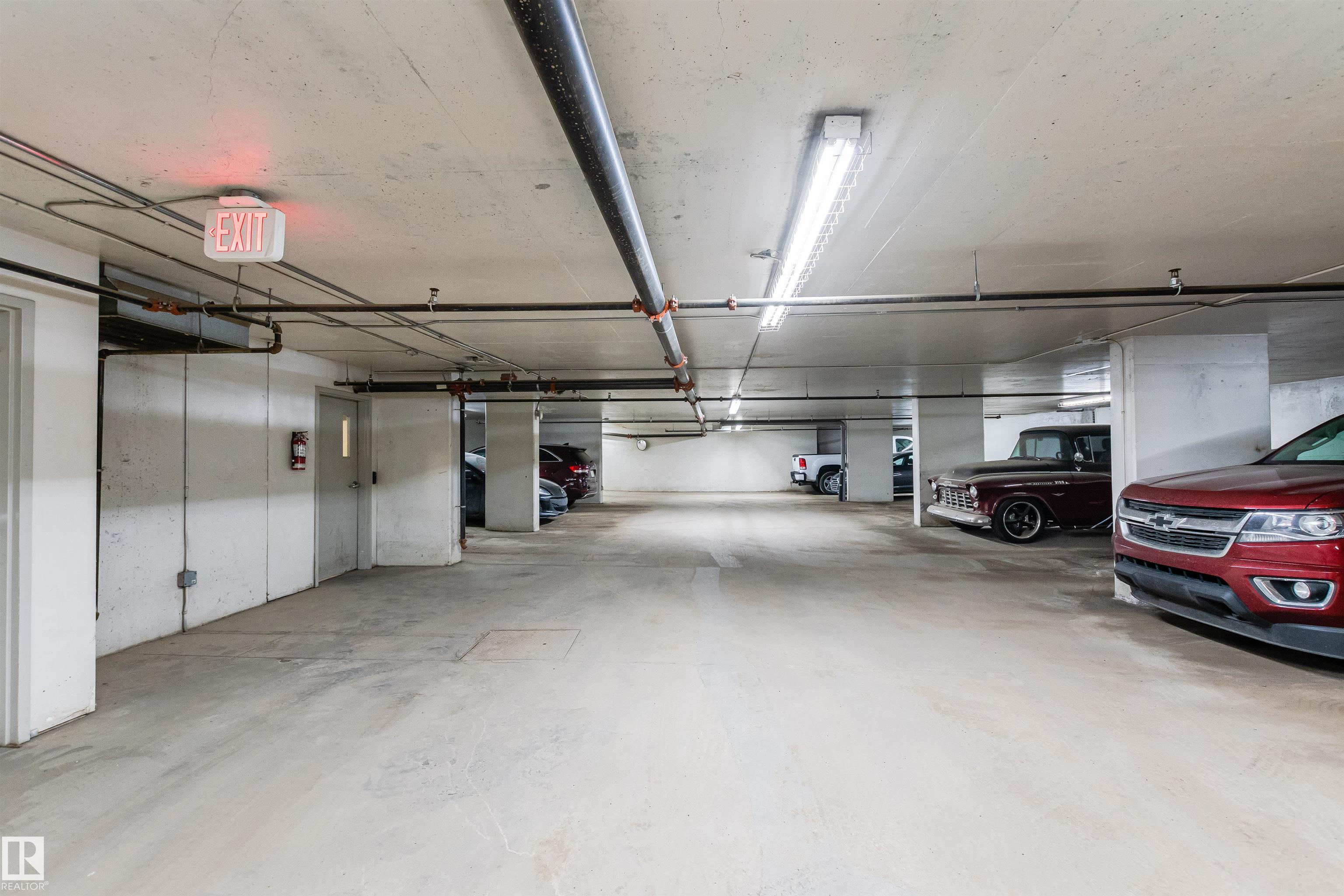 Underground parking garage with concrete pillars, overhead pipes, and parked vehicles.