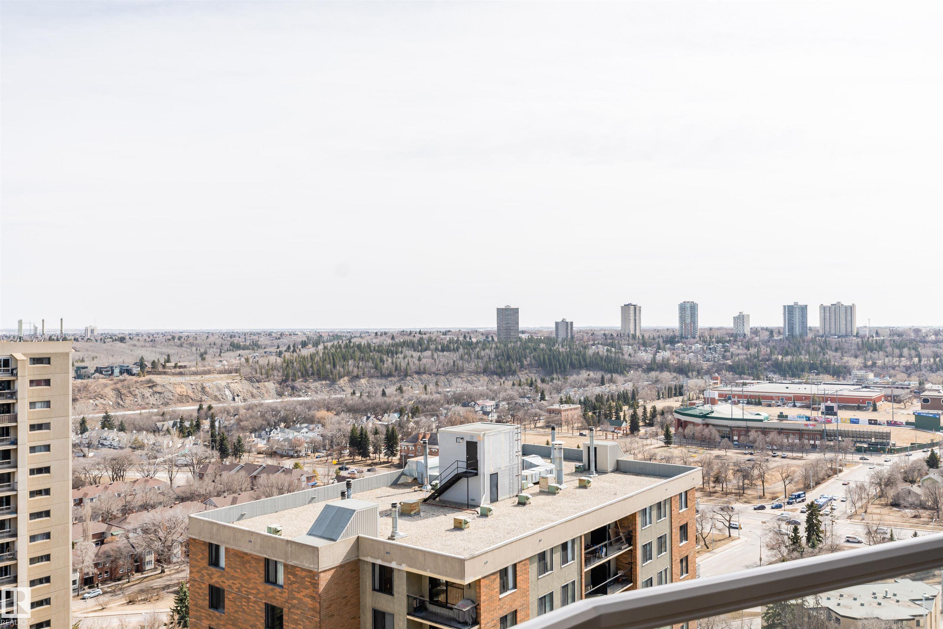 Panoramic Edmonton city view from a high-rise balcony, with a wooded ravine and distant towers.