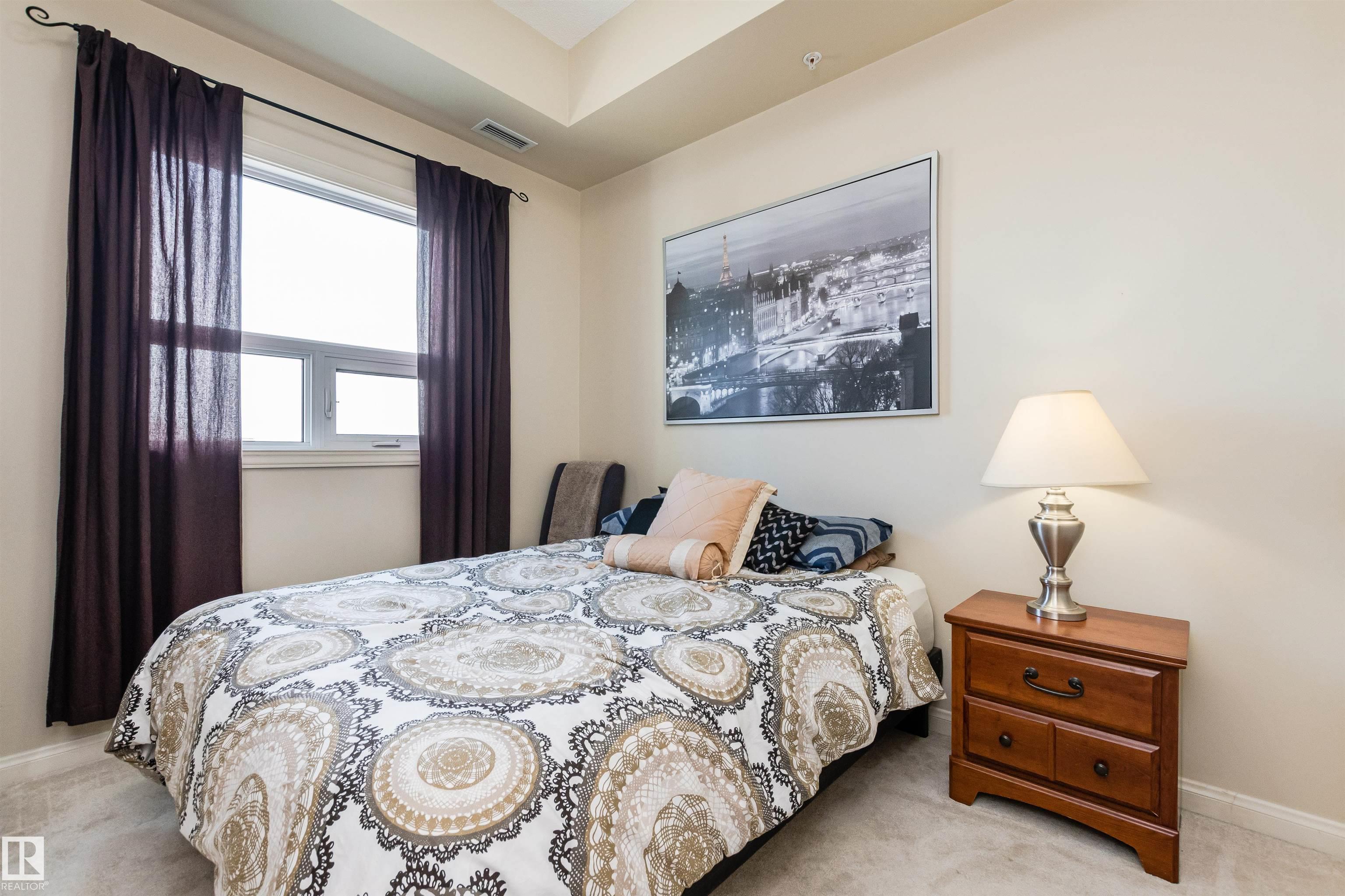 Furnished bedroom with a patterned bedspread, dark curtains, and a framed Paris cityscape print.