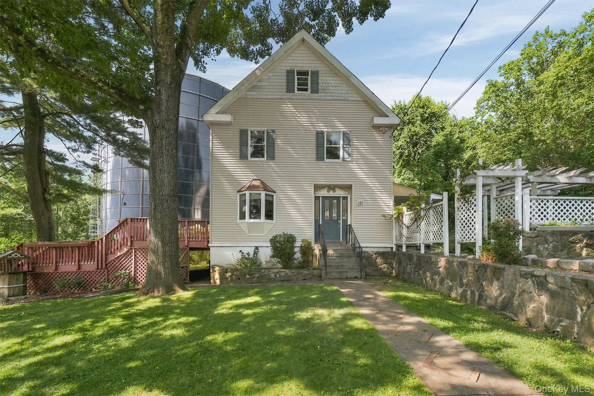 Two-story gable-front house with a bay window, red deck, white pergola, and stone retaining wall.