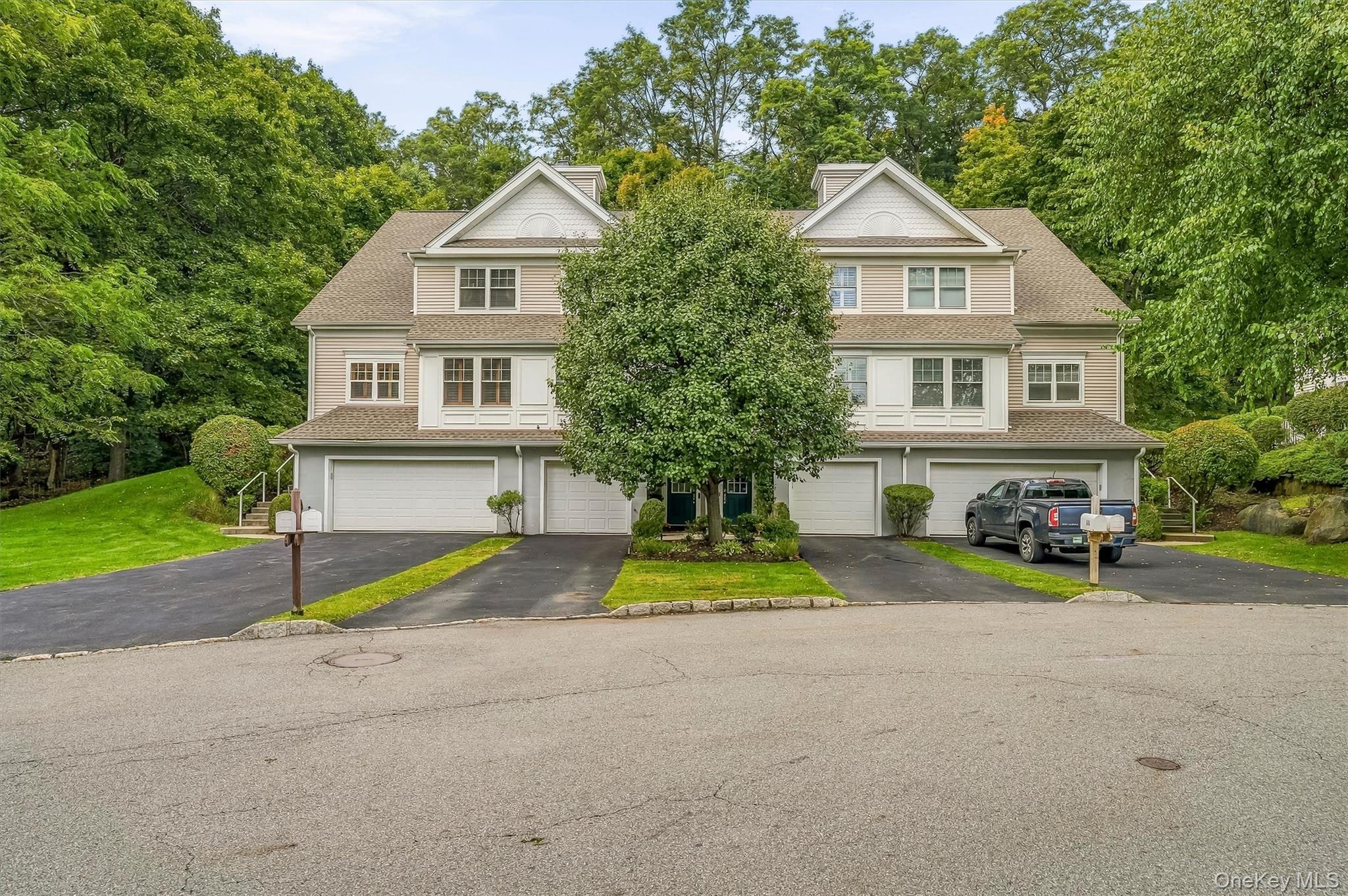 Attached beige residence with multiple garages, a central tree, and wooded surroundings.