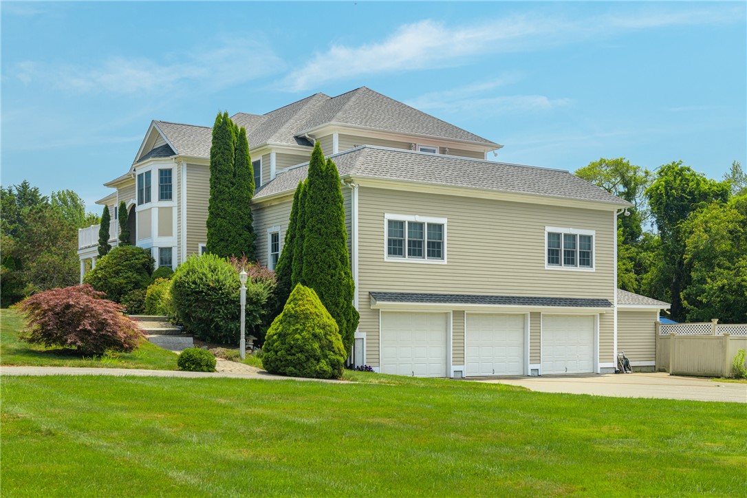 Two-story beige house with a three-car garage, landscaped shrubs, and a broad green lawn.