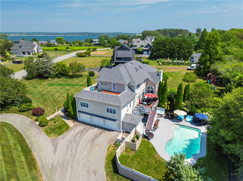 Aerial view of a gray-shingled home with a swimming pool, patio, landscaped yard, and water in the distance.