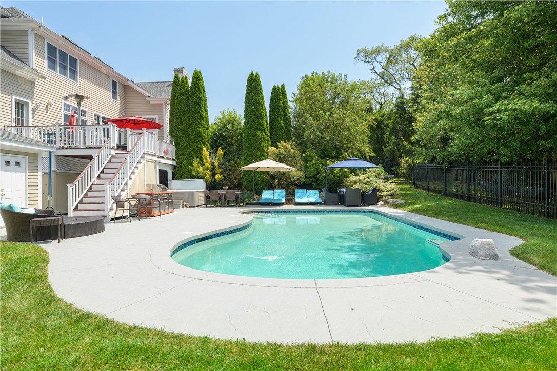 Curved backyard swimming pool with a broad patio, raised deck, and tree-lined fenced lawn.