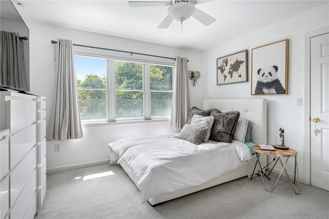 Bright bedroom with a white bed, wide window, leafy outdoor view, and framed wall art.