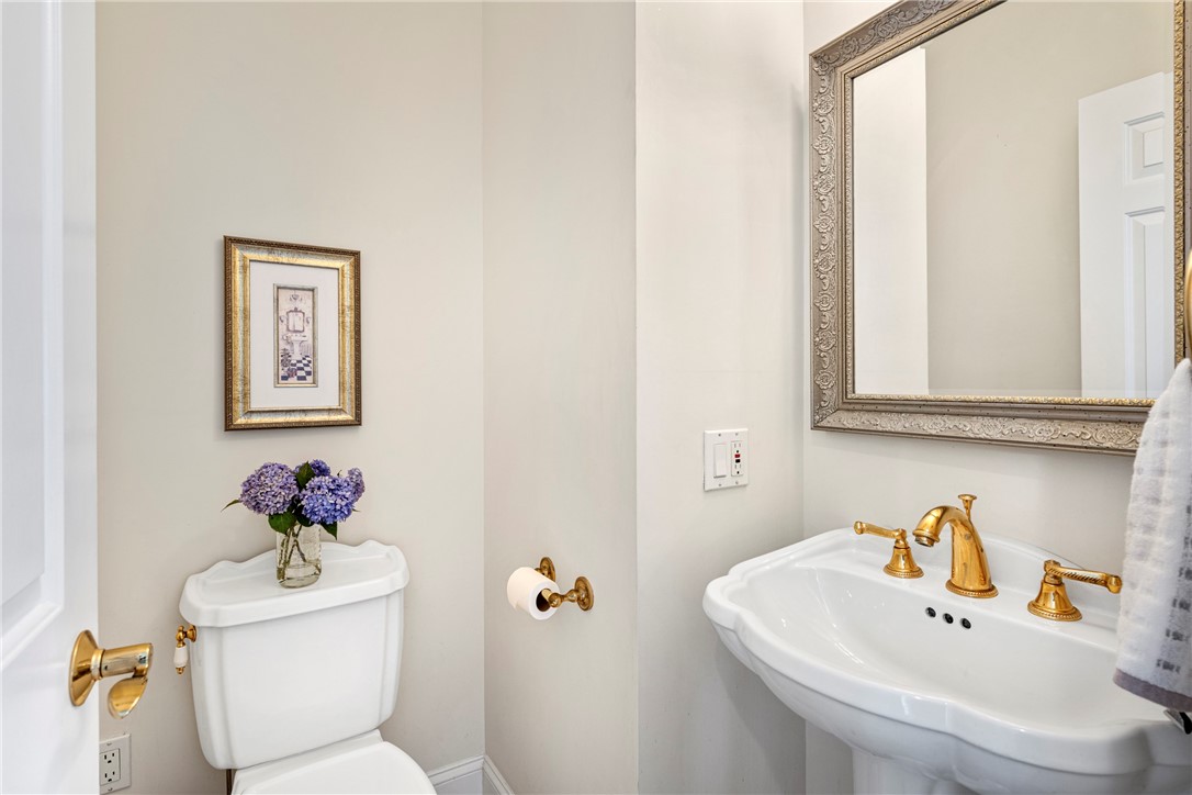 Powder room with a curved white sink, gold fixtures, ornate mirror, and purple flowers on the toilet tank.