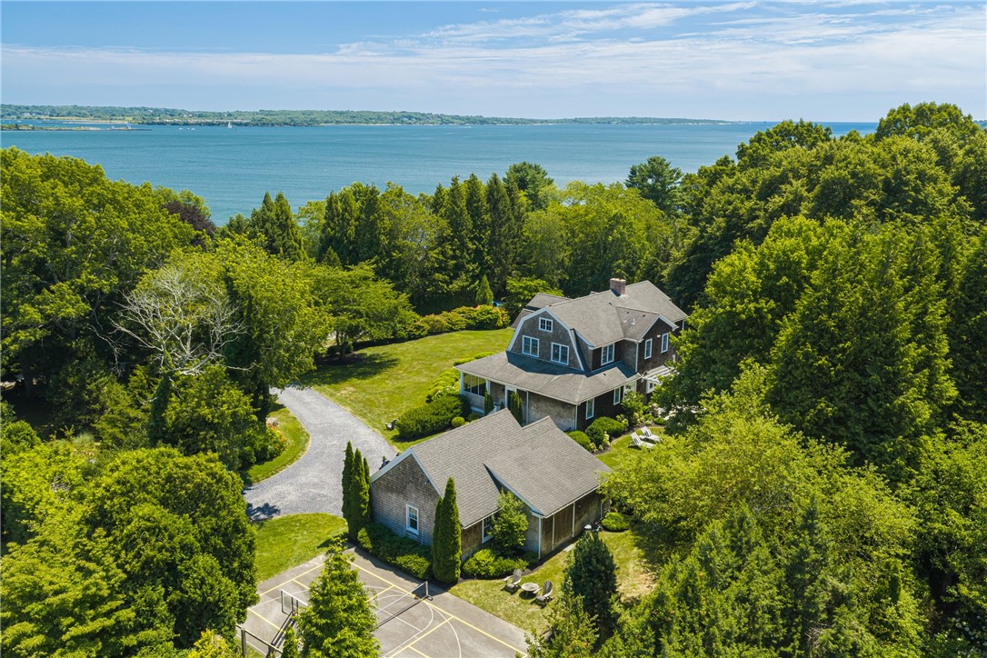 Aerial view of a shingled home surrounded by trees, with a sport court and water beyond.