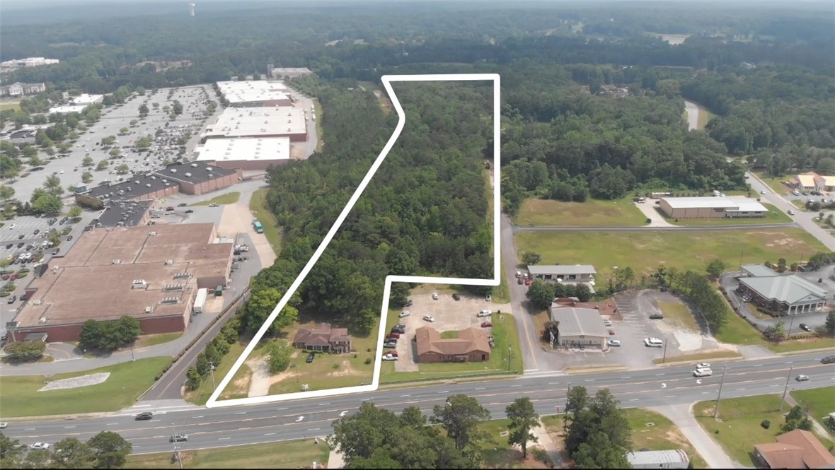 Wooded land parcel outlined in white beside a multi-lane road and commercial buildings.