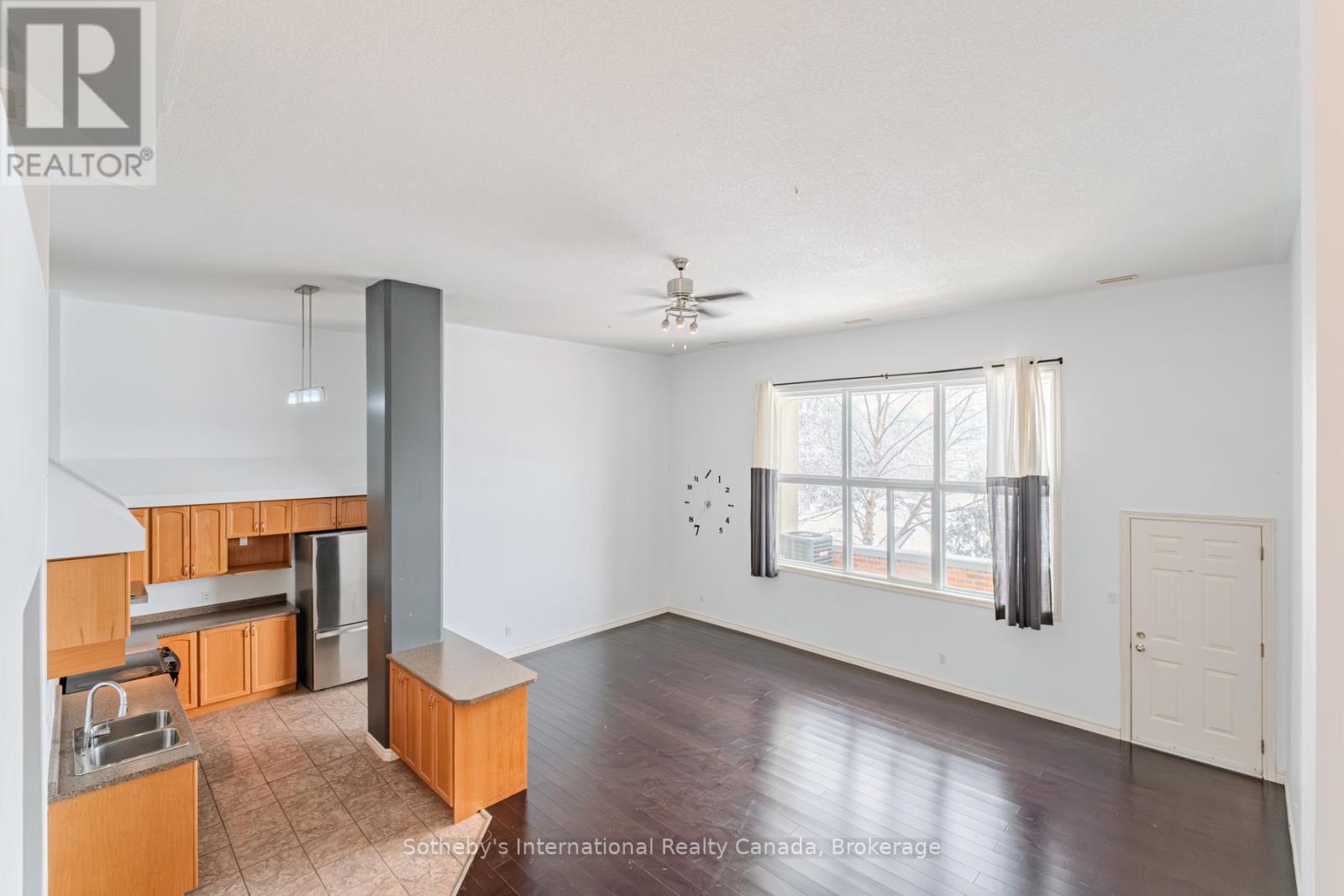 Open living room and kitchen with dark hardwood floors, warm wood cabinets, and a large curtained window.