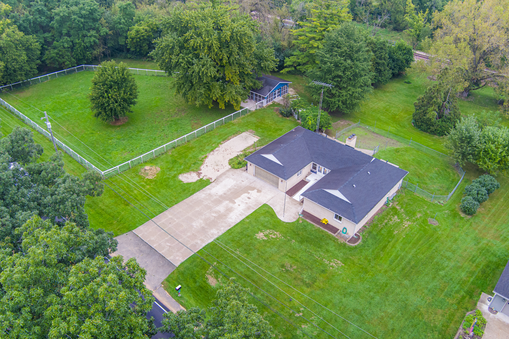 Aerial view of a house with a broad driveway, fenced lawn, and detached outbuilding.