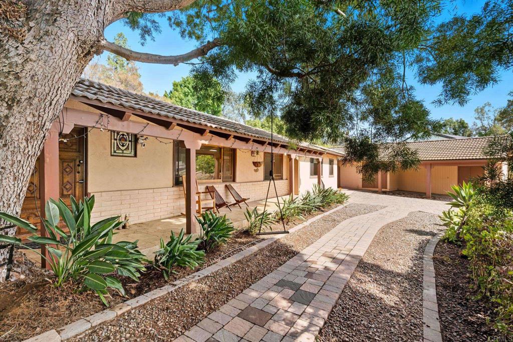 Single-story home with a covered patio, outdoor seating, and a paved walkway beneath a large tree.